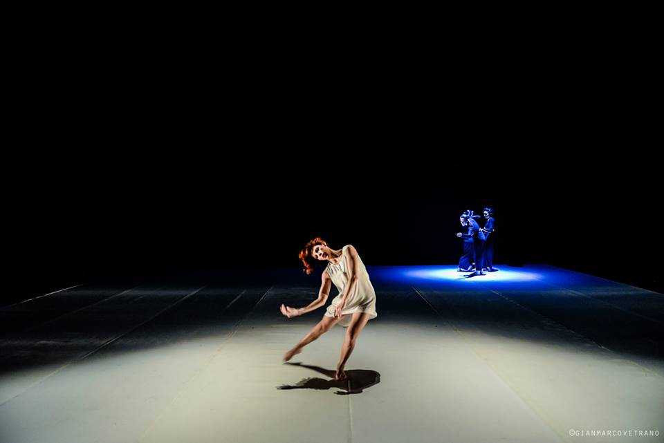 Scena di danza illuminata: una figura in un fascio di luce e due persone in controluce sullo sfondo